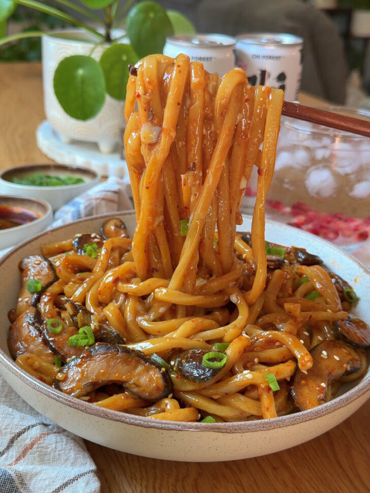 Chopsticks lifting glossy spicy garlic butter udon noodles with shiitake mushrooms, scallions, and sesame seeds.