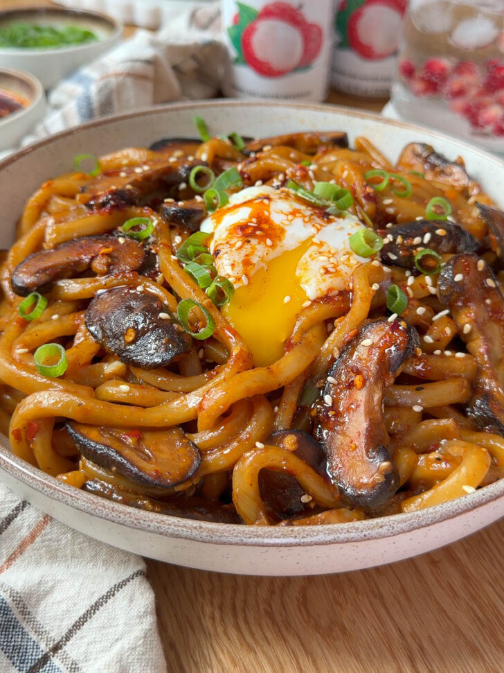 Bowl of glossy spicy garlic butter udon topped with shiitake mushrooms, a runny onsen egg, scallions, and sesame seeds.